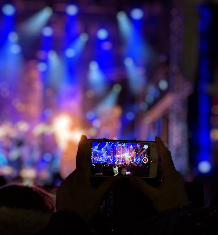 Home Silhouetted hands holding a smartphone capturing vibrant concert lights.